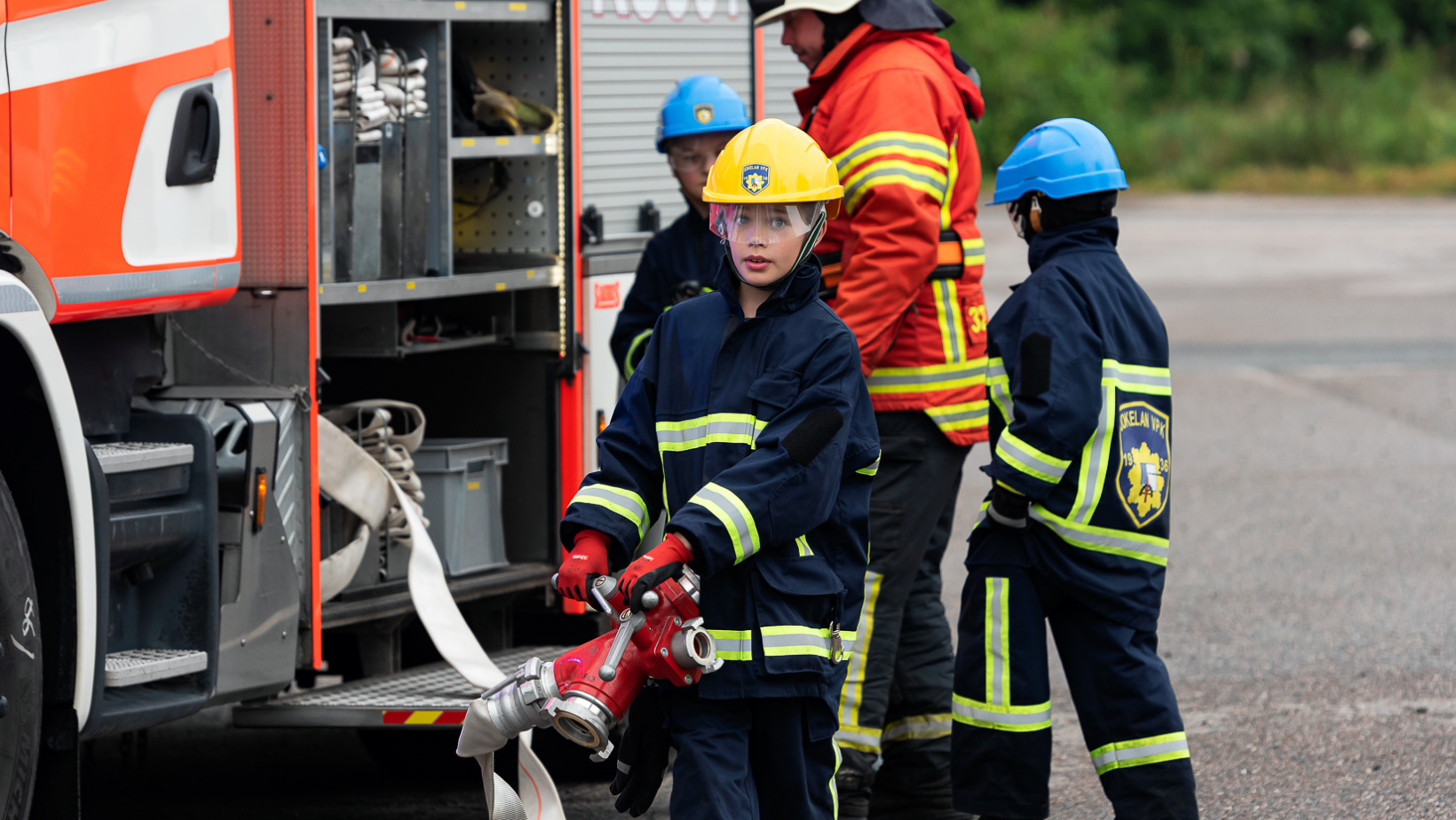 Kuvassa kolme palokuntanuorta vetämässä letkua paloautosta. Kuvassa myös kouluttaja ohjaamassa toimintaa.